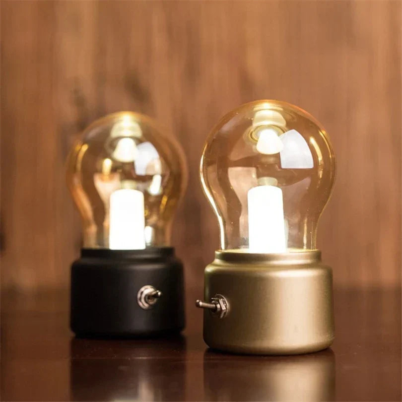 Two light bulb-shaped lamps, one black and one gold, on a wooden surface with a blurred wooden background.