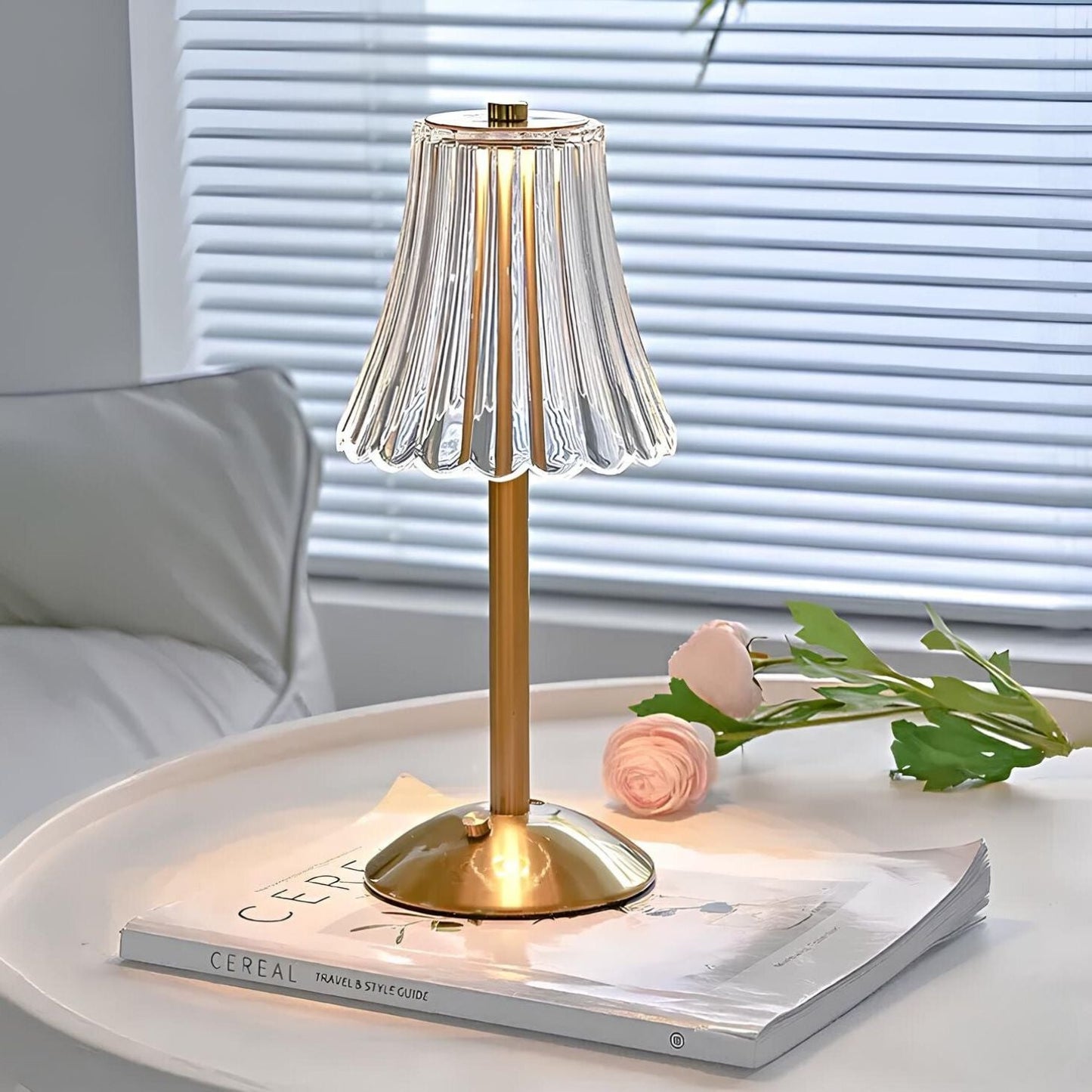 Gold table lamp with a pleated glass shade on a surface with a magazine and flowers.