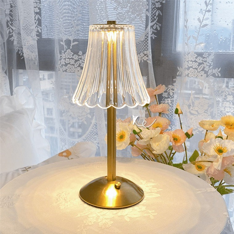 Gold table lamp with a clear glass shade on a surface with flowers and lace curtains in the background.