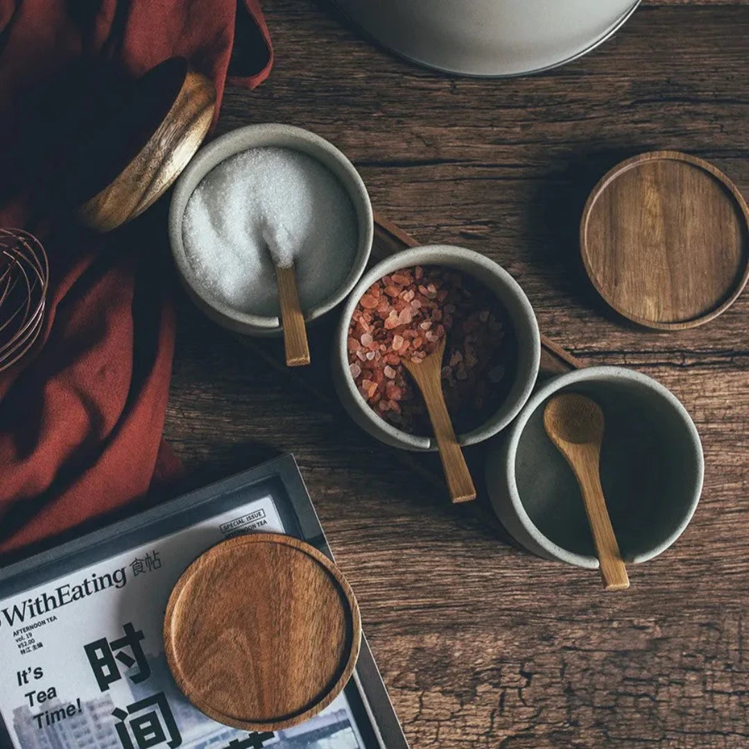 Josh & Tate - Ceramic Condiment Storage Set - Three small bowls with wooden spoons on a wooden table, with a magazine partially visible.