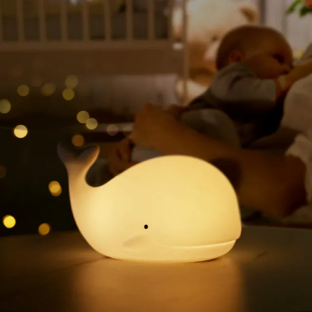 Whale-shaped night light on a surface with blurred background of a child and teddy bear.