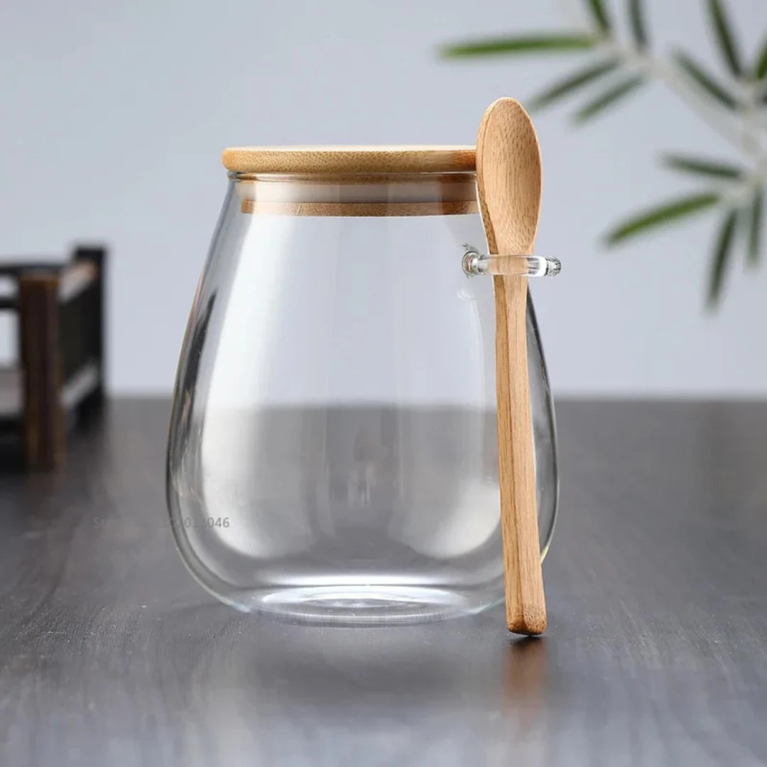 Josh & Tate - Glass Storage Jar with Spoon - Clear glass jar with wooden lid and spoon on a gray surface