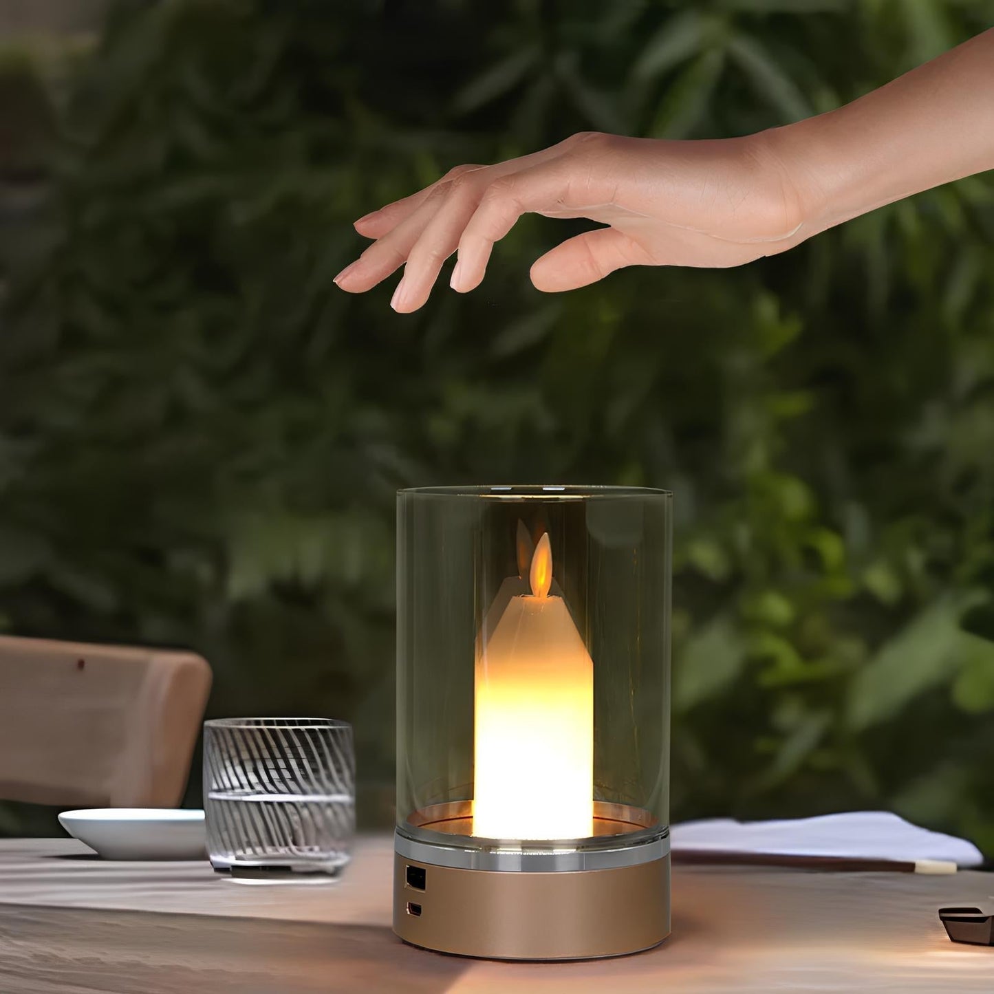 Glass candle holder with flame on a table, hand reaching towards it, blurred greenery in the background