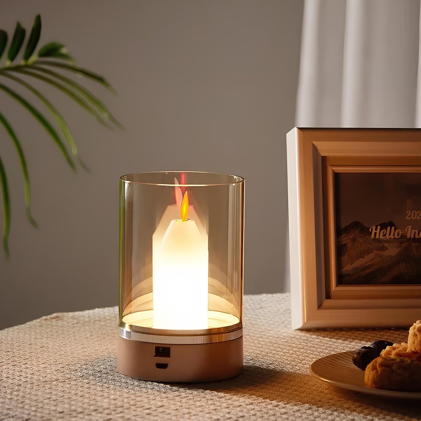 Small lamp with a candle-like light on a surface next to a photo frame and plant.