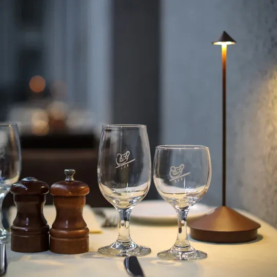 Two wine glasses with a logo on a table next to salt and pepper shakers, with a blurred background.