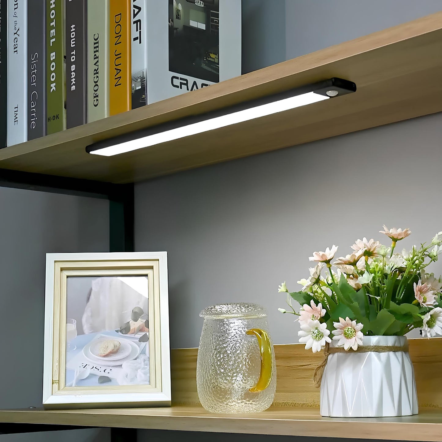 Wooden shelf with books, a picture frame, a glass pitcher, and a vase with flowers against a gray wall.