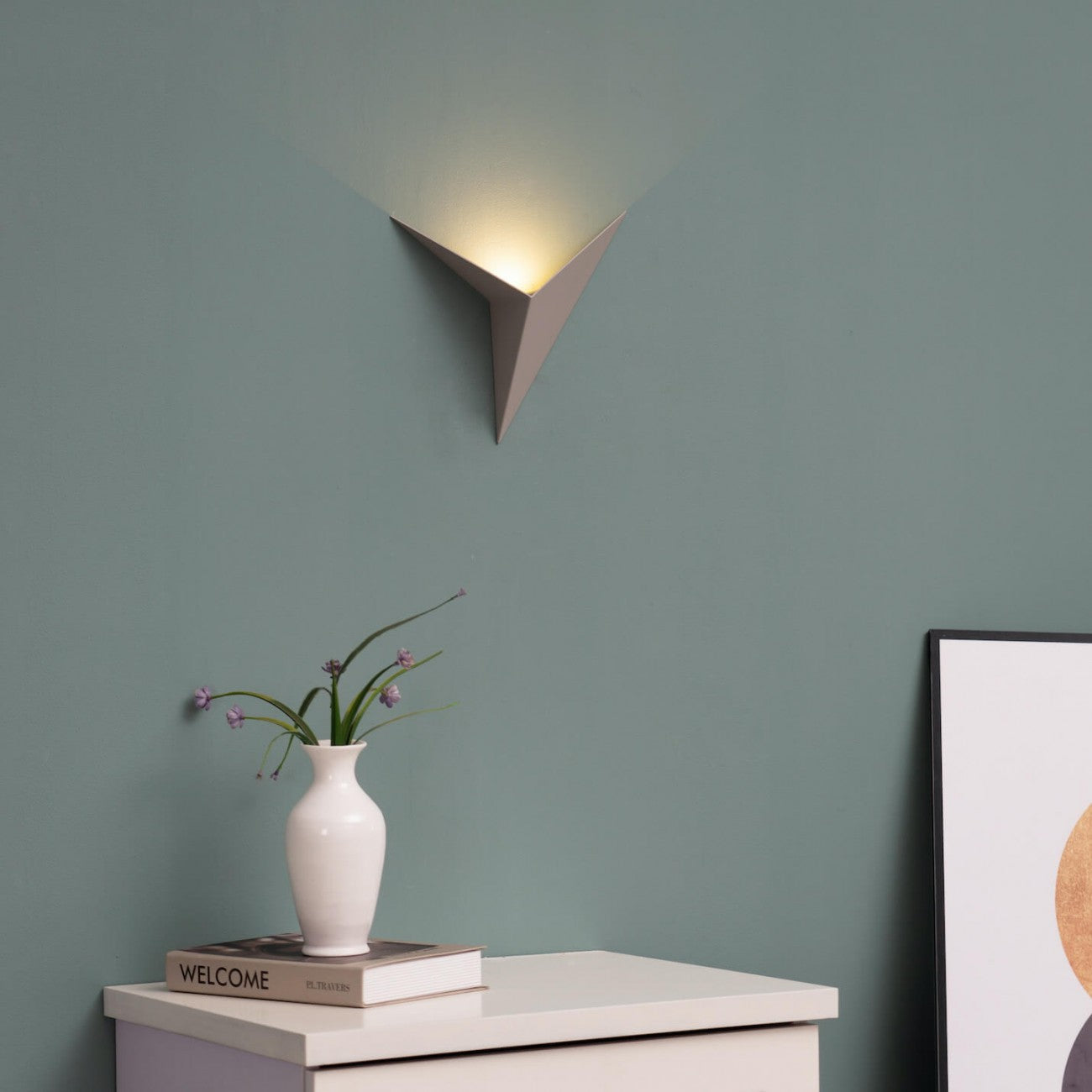Wall-mounted lamp casting light on a table with a vase and books against a green wall.