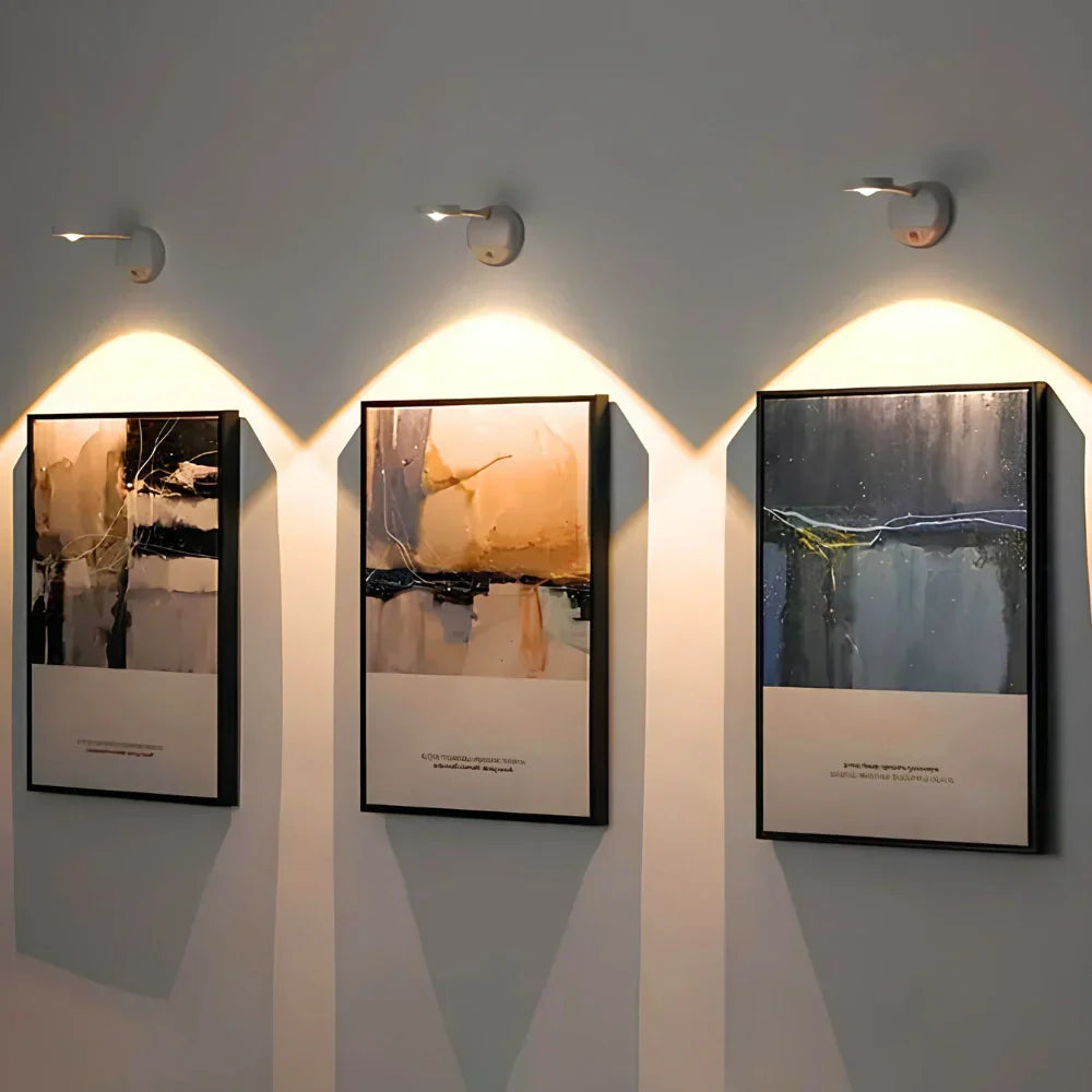 Three framed artworks on a wall with wall lights illuminating them.