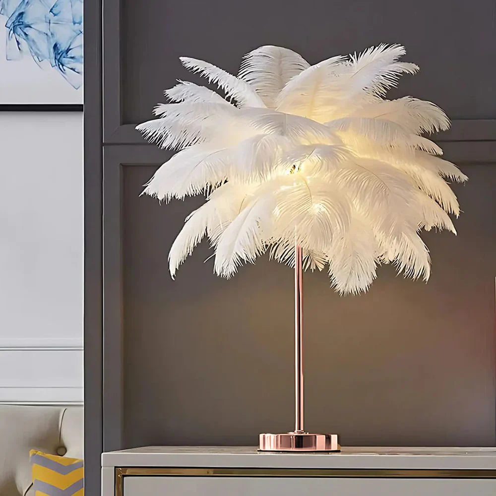 Feathered table lamp with a rose gold base on a shelf against a gray wall.