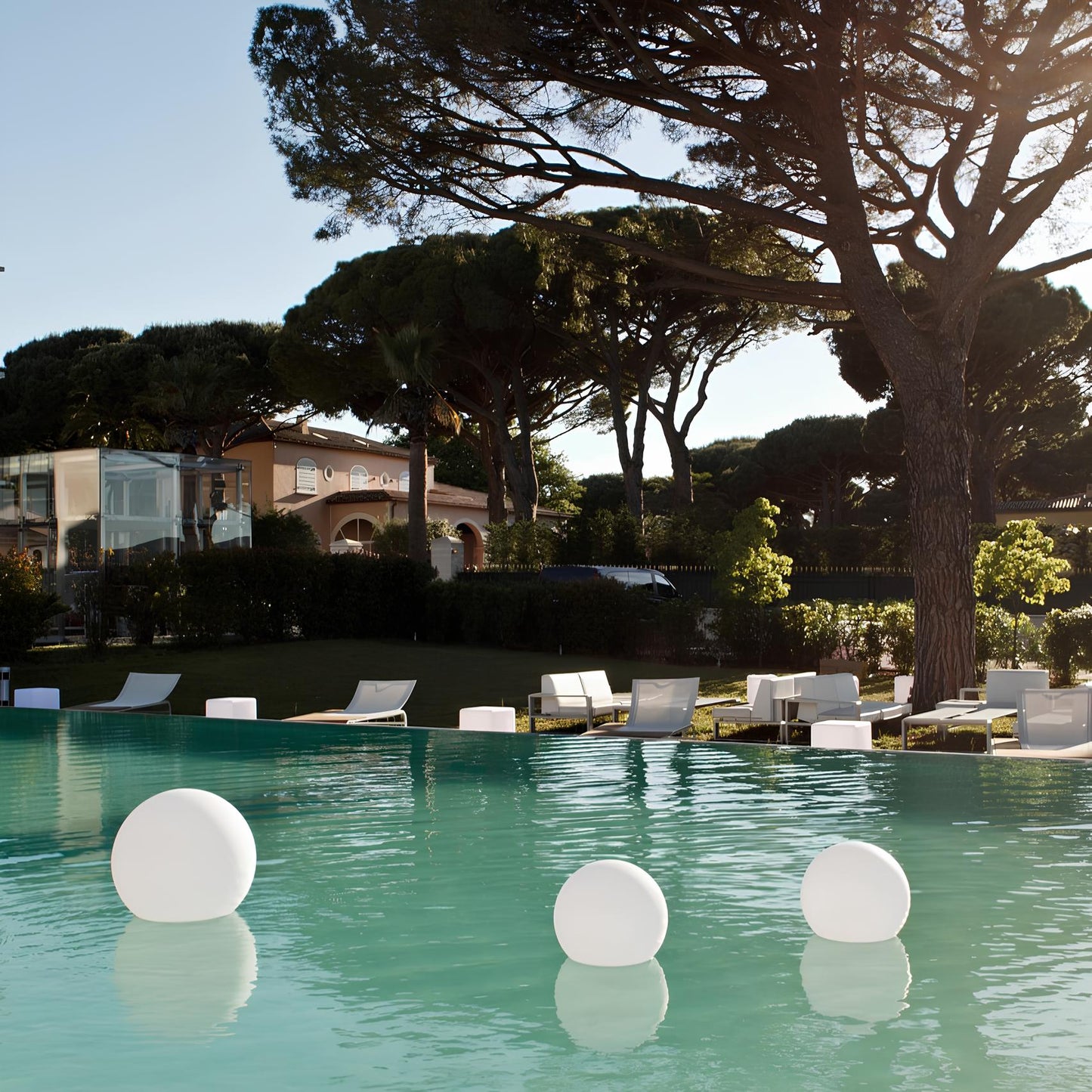 Pool area with lounge chairs and floating lights, surrounded by trees and buildings.