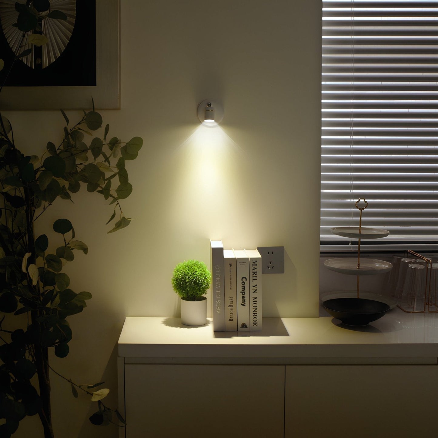 Wall-mounted light fixture illuminating a small table with books and a plant in a dimly lit room.