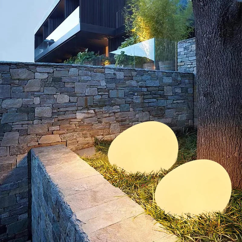 Two spherical outdoor lights on grass next to a stone wall with modern building in the background