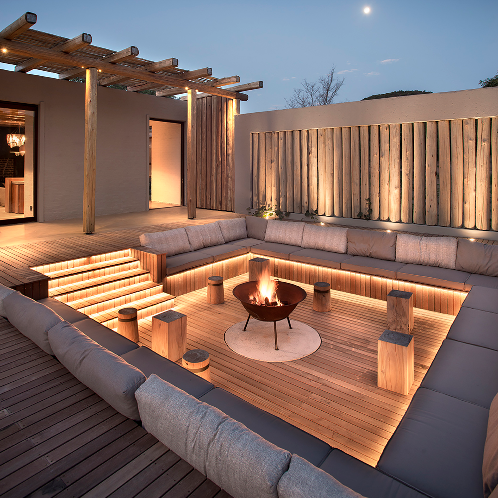Modern outdoor patio with seating area, fire pit, and wooden pergola at night.
