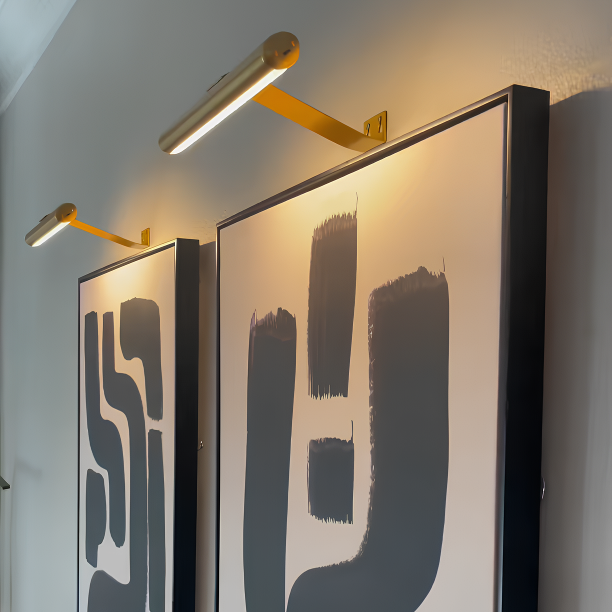 Two abstract art pieces with black brush strokes on a white background, illuminated by gold wall lights.