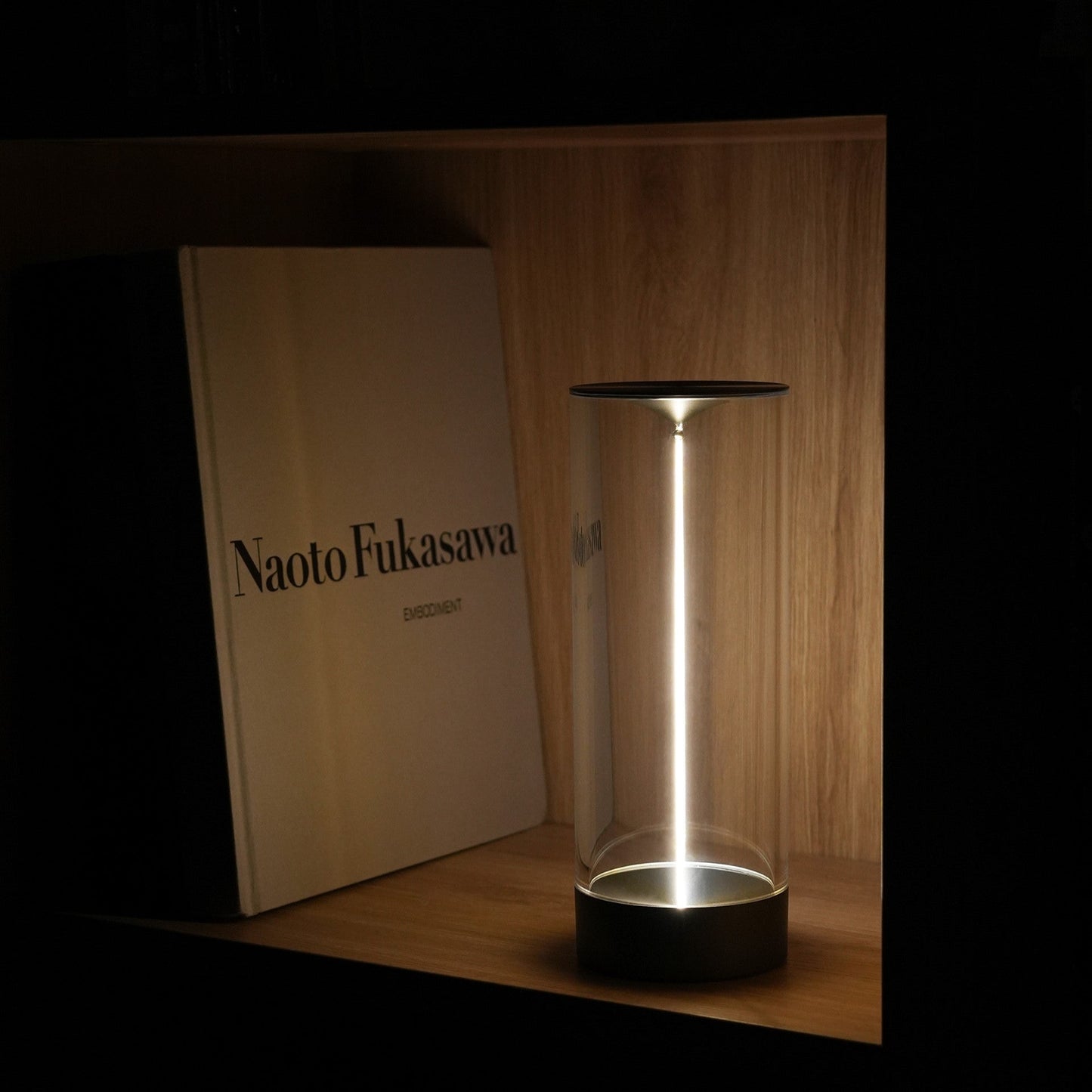 Clear cylindrical lamp with black base on a wooden surface, with 'Naoto Fukasawa' book in the background.