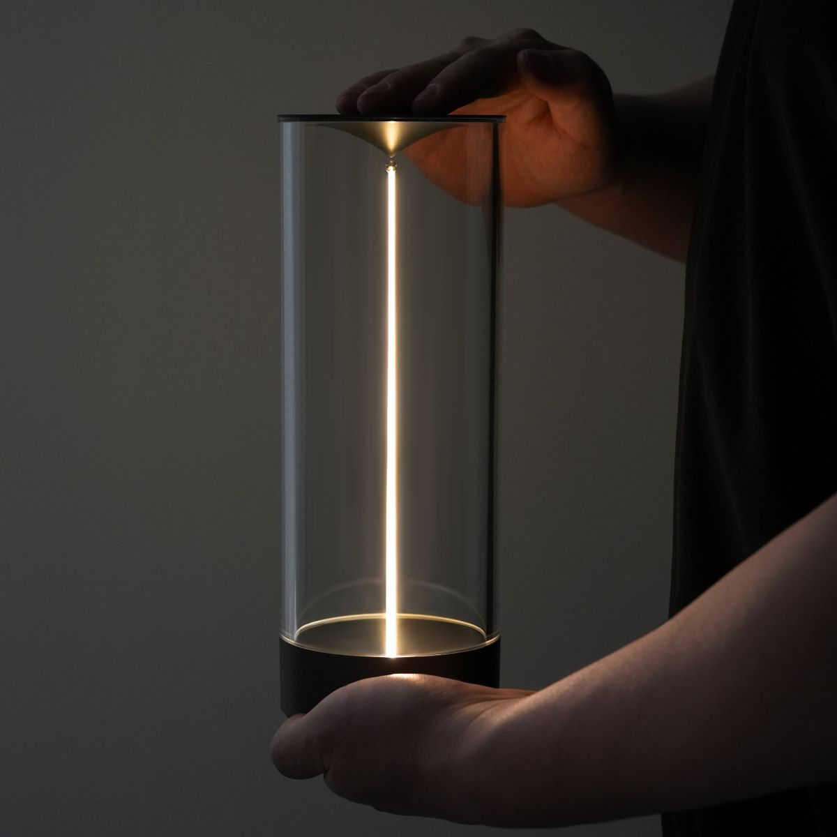 Person holding a cylindrical light fixture with a glowing interior against a dark background