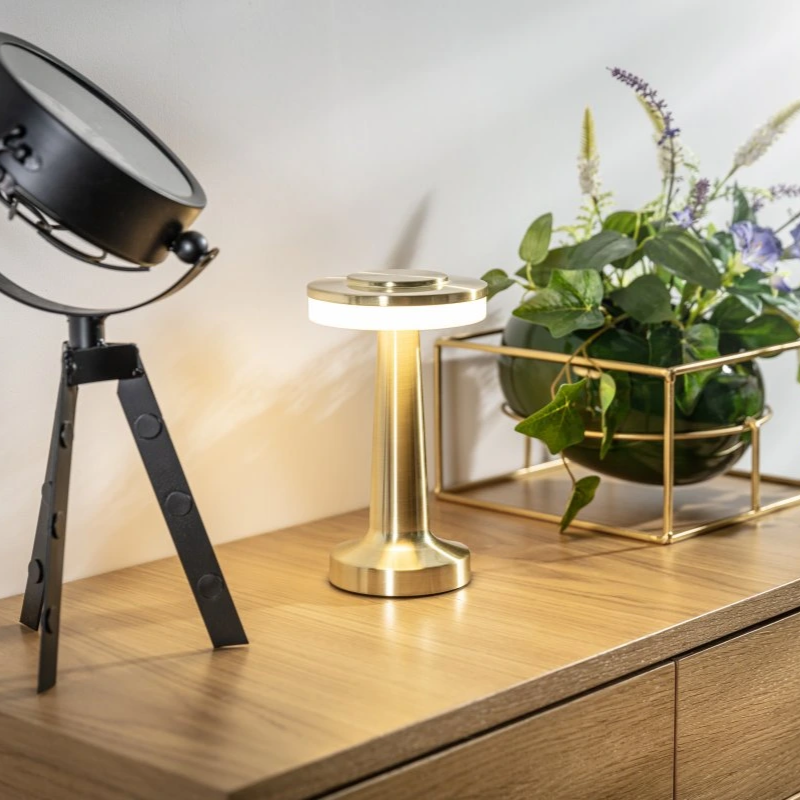 Gold table lamp on a wooden surface with a black lamp and plant in the background