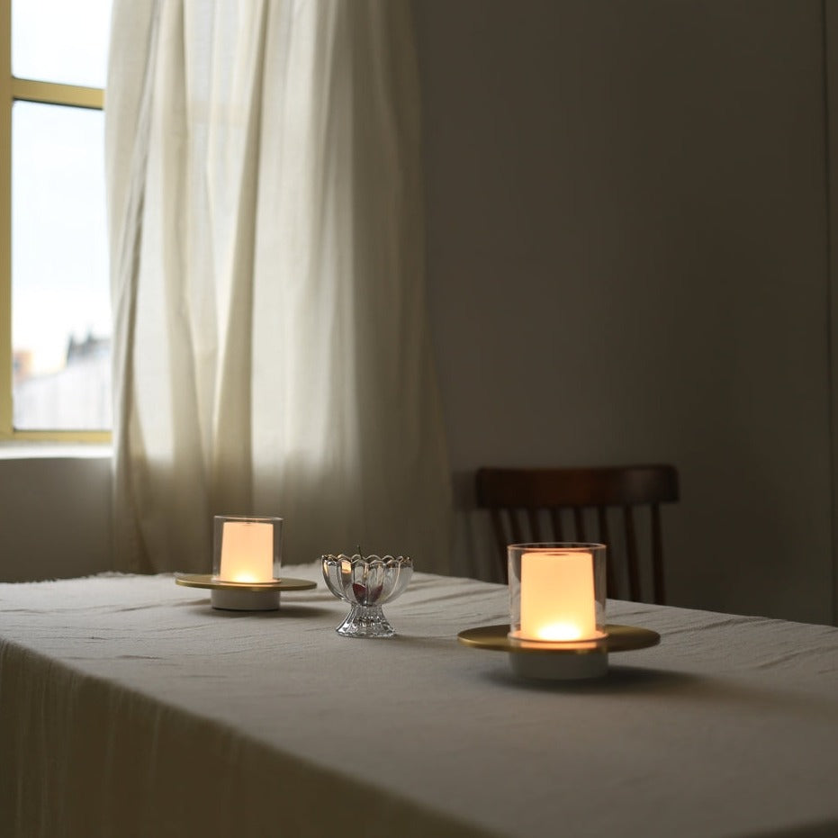 Two lit candles on a table with a window and chair in the background