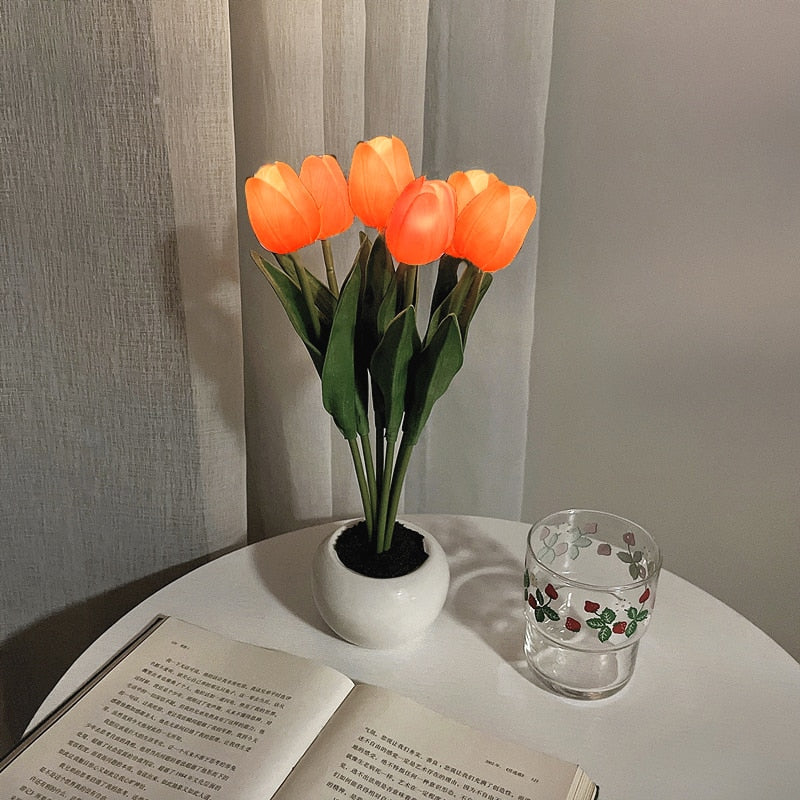 Vase with glowing orange tulips on a table next to an open book and a glass.