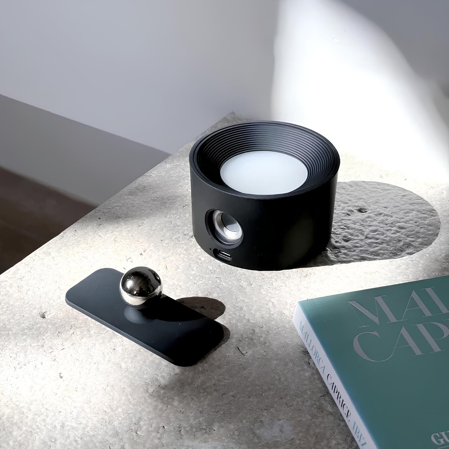 Black cylindrical object with a white candle inside on a concrete surface next to a book.