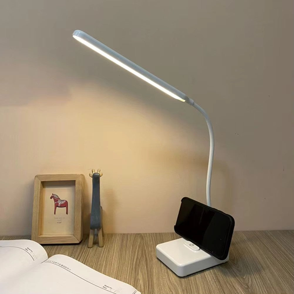 Desk lamp with phone holder on a wooden surface next to a book and small figurine.