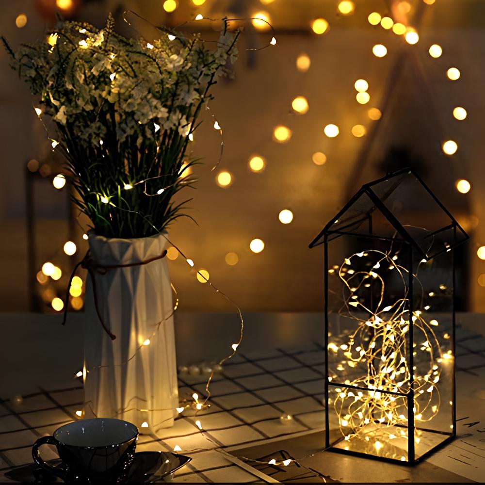 Decorative setup with a vase of flowers, lit lantern, and cup on a table with string lights in the background.