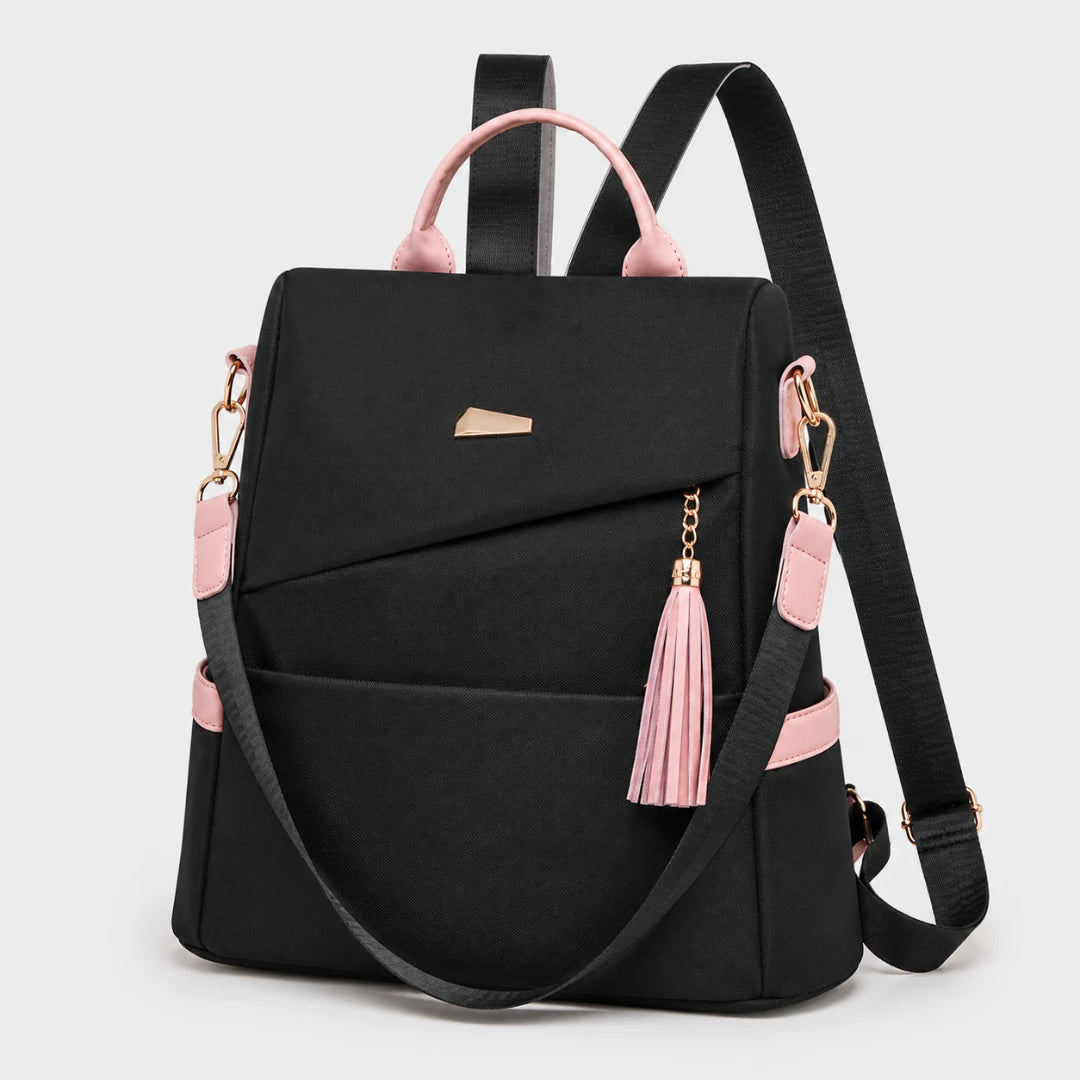 Josh & Tate - AntiTheft™ Oxford Backpack - A black backpack with light pink accents, including the top handles, zipper pulls, and side details, featuring gold-tone hardware and a matching pink tassel charm, shown on a light gray background.