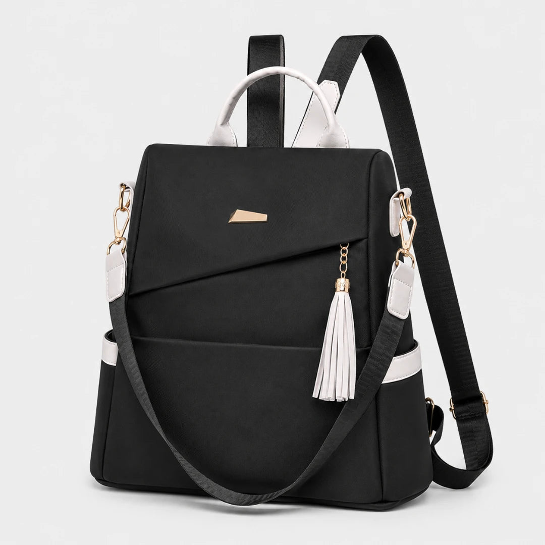 Josh & Tate - AntiTheft™ Oxford Backpack - A black backpack with white accents, featuring a top handle, adjustable shoulder strap, a hanging white tassel charm, and gold-tone hardware, shown on a light gray background.