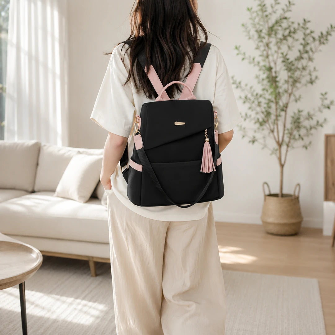 Josh & Tate - AntiTheft™ Oxford Backpack - A woman viewed from behind wearing a black backpack with light pink straps, tassel detail, and a small gold logo on a light-colored sofa in a bright living room.