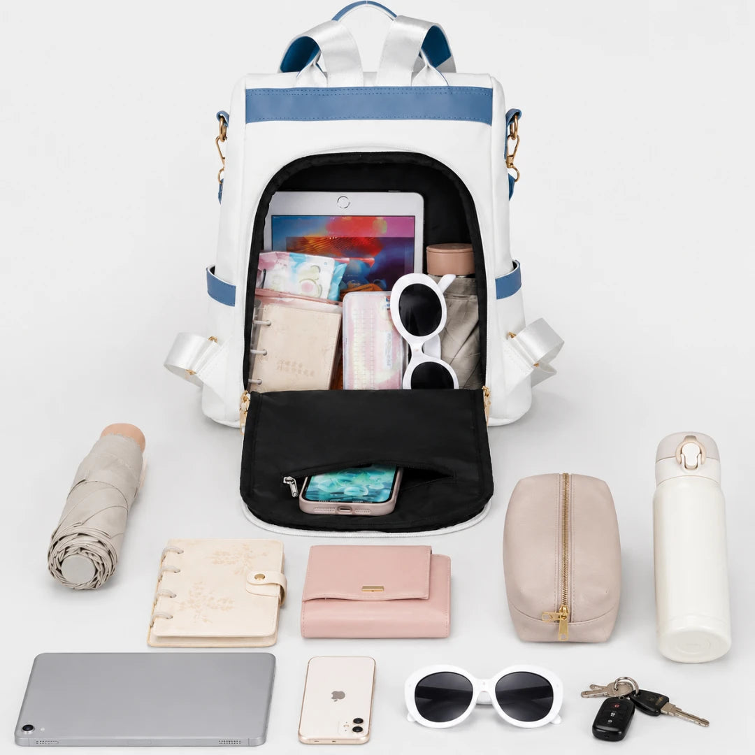 Josh & Tate - AntiTheft™ Oxford Backpack - A white backpack with blue trim and gold-tone hardware is shown open, filled with various accessories including a tablet, sunglasses, wallet, phone, and small bags. Surrounding the backpack on a clean white background are matching accessories: a rolled beige umbrella, a cream notebook, a pink wallet, a beige pouch, a white thermos, a tablet, a phone, white sunglasses, and keys.