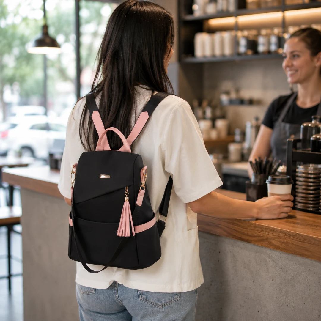 Josh & Tate - AntiTheft™ Oxford Backpack - A woman seen from behind wearing a black backpack with pink straps, zipper pulls, and tassel details while standing at a cafe counter with a barista behind her.