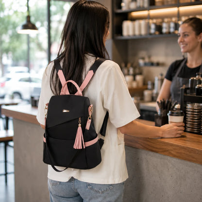 Josh & Tate - AntiTheft™ Oxford Backpack - A woman seen from behind wearing a black backpack with pink straps, zipper pulls, and tassel details while standing at a cafe counter with a barista behind her.