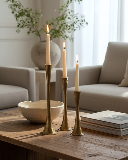 Three lit candles in gold candle holders on a wooden table with a cozy living room background.