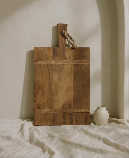 Josh & Tate - Archer Wood Cutting Board - Wooden cutting board with a handle on a light surface with a vase in the background