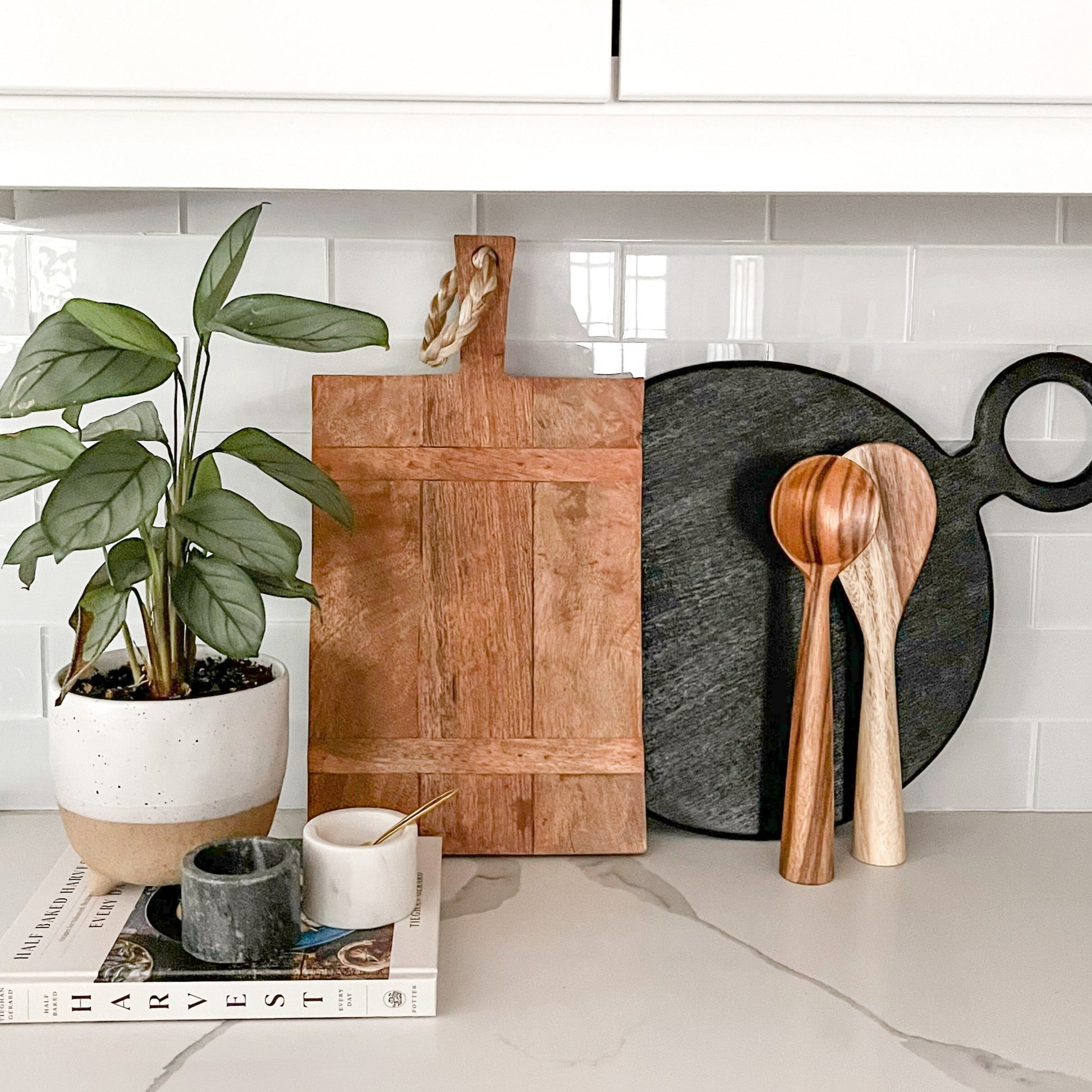 Wooden cutting board, black cutting board, and wooden spoons on a kitchen counter with a plant and books in the background.