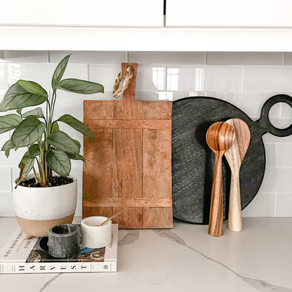 Wooden cutting board, black cutting board, and wooden spoons on a kitchen counter with a plant and books in the background.