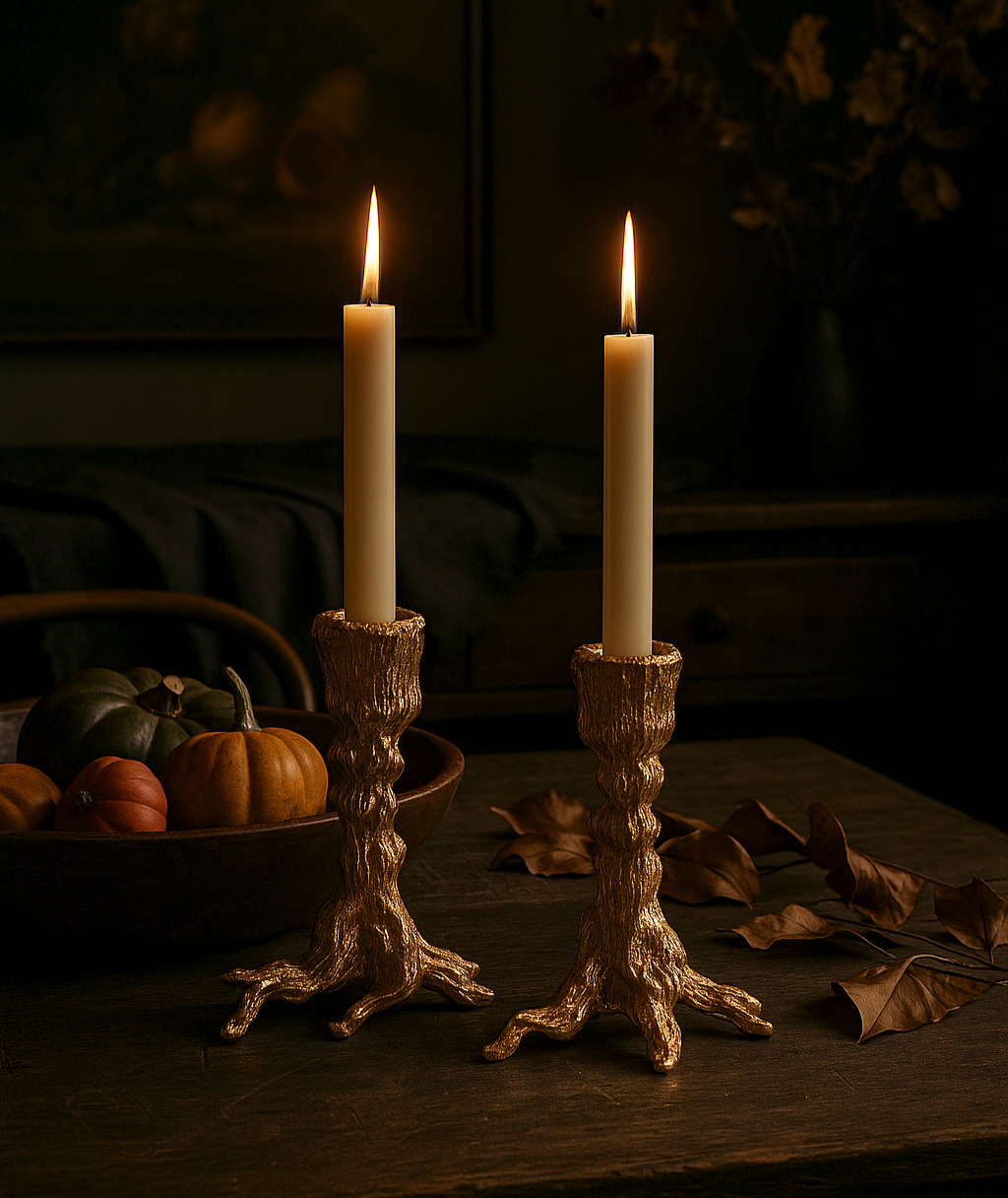 Two lit candles in gold candlesticks on a dark surface with pumpkins and leaves.