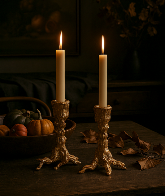 Two lit candles in gold candlesticks on a dark surface with pumpkins and leaves.