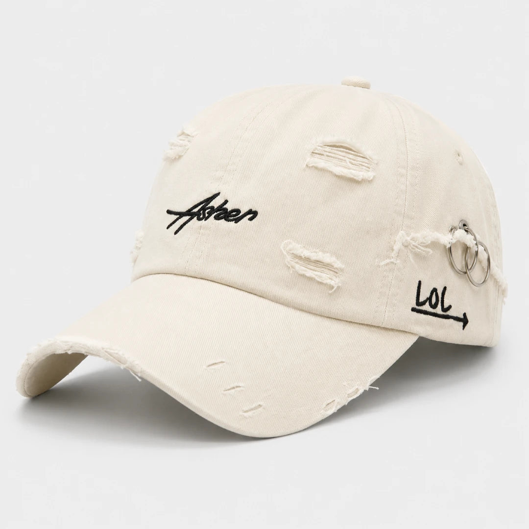 Josh & Tate - AsherLOL™ Distressed Cap - A distressed off-white baseball cap with black embroidered text reading 'Aster' on the front and 'LOL' with an arrow on the side, featuring metal ring details and distressed rips on the brim and crown.