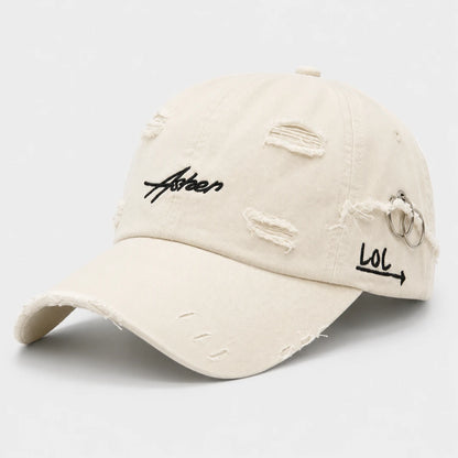 Josh & Tate - AsherLOL™ Distressed Cap - A distressed off-white baseball cap with black embroidered text reading 'Aster' on the front and 'LOL' with an arrow on the side, featuring metal ring details and distressed rips on the brim and crown.