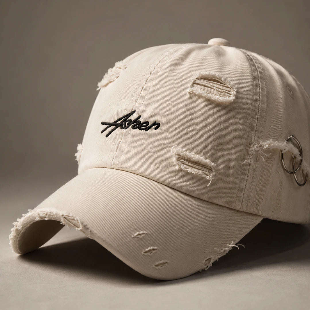 Josh & Tate - AsherLOL™ Distressed Cap - A distressed off-white baseball cap with a black embroidered 'Aster' logo on the front, featuring frayed details and a silver metal ring accent on the side, set against a neutral gray background.