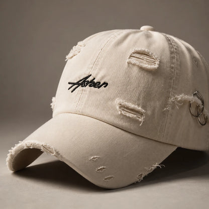 Josh & Tate - AsherLOL™ Distressed Cap - A distressed off-white baseball cap with a black embroidered 'Aster' logo on the front, featuring frayed details and a silver metal ring accent on the side, set against a neutral gray background.
