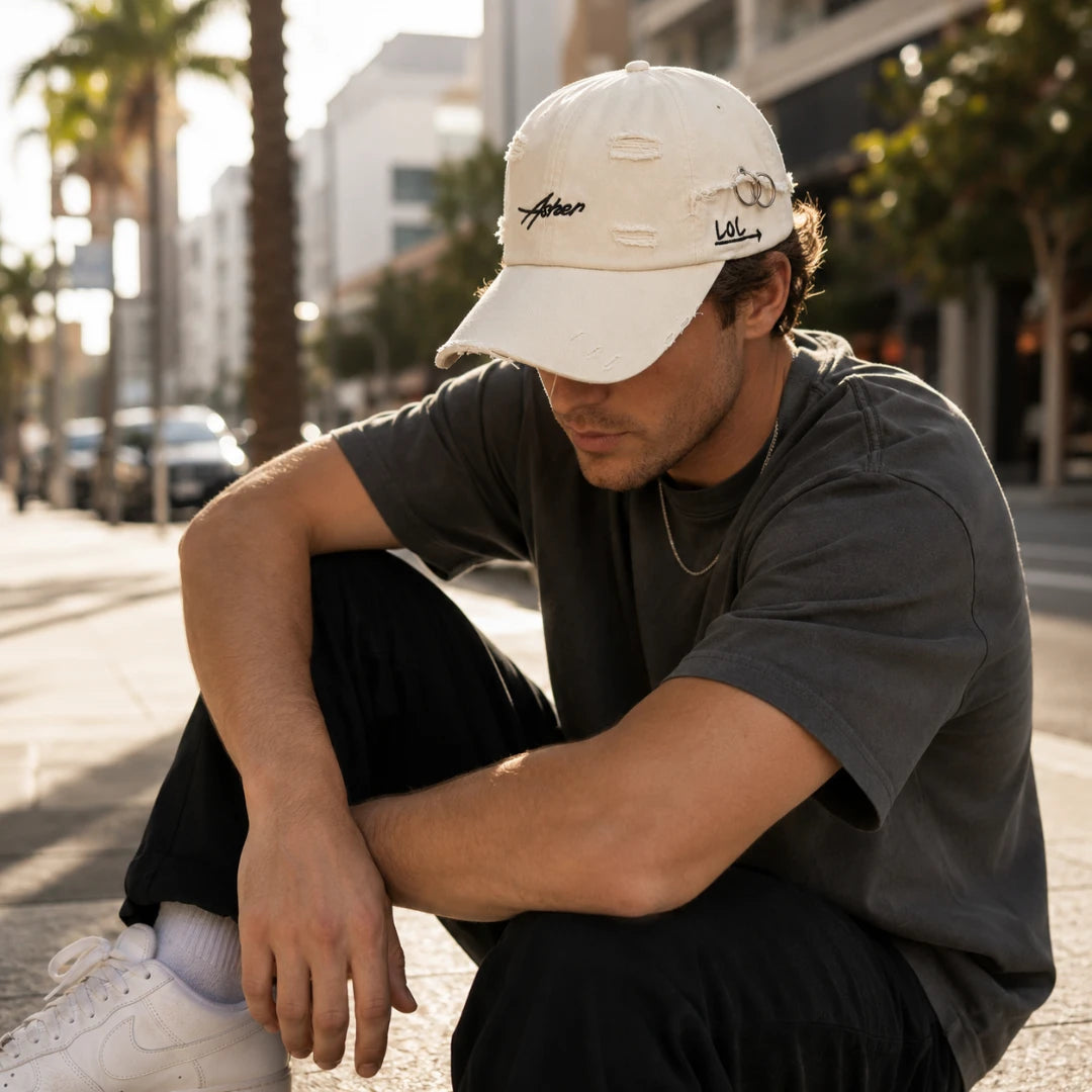 Josh & Tate - AsherLOL™ Distressed Cap - A man crouching on a city sidewalk wearing a distressed off-white baseball cap with embroidered script logos, a dark gray t-shirt, black pants, and white sneakers. The background shows a blurred urban street scene with palm trees.
