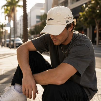 Josh & Tate - AsherLOL™ Distressed Cap - A man crouching on a city sidewalk wearing a distressed off-white baseball cap with embroidered script logos, a dark gray t-shirt, black pants, and white sneakers. The background shows a blurred urban street scene with palm trees.