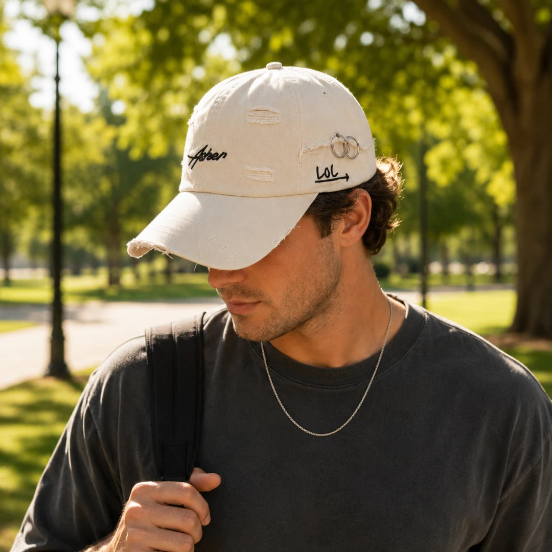 Josh & Tate - AsherLOL™ Distressed Cap - A man wearing a distressed off-white baseball cap with embroidered script details, photographed outdoors in a sunny park with blurred green trees in the background.