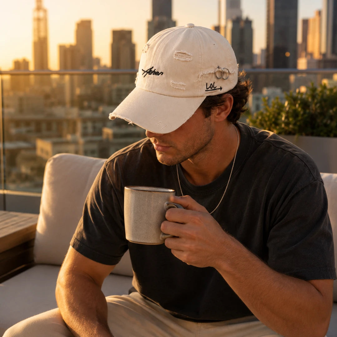 Josh & Tate - AsherLOL™ Distressed Cap - A man sitting on a patio at sunset wearing a distressed off-white baseball cap with embroidered script logos, a dark gray t-shirt, and holding a metal mug. The background shows a blurred city skyline.