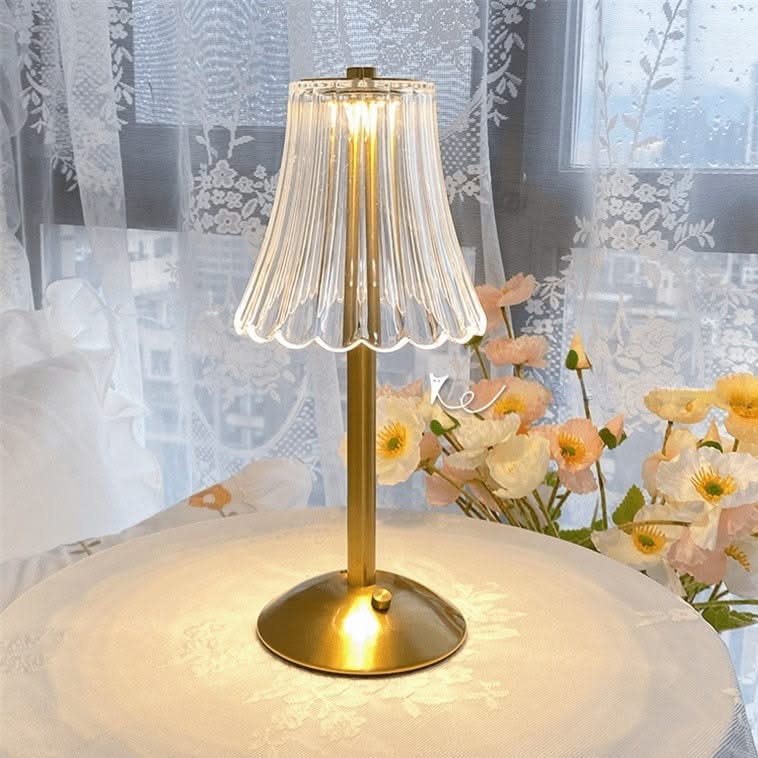 Gold table lamp with a clear glass shade on a surface with flowers and lace curtains in the background.