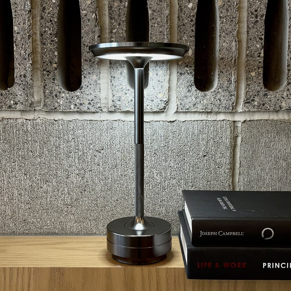 Black desk lamp on a wooden surface with a book titled 'Life & Work' by Joseph Campbell against a stone wall background.