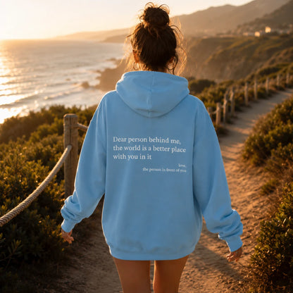Josh & Tate - BehindMe™ Quote Hoodie - A woman viewed from behind wearing a light blue hoodie with a white printed quote on the back, standing on a sandy path overlooking a coastal ocean at sunset.