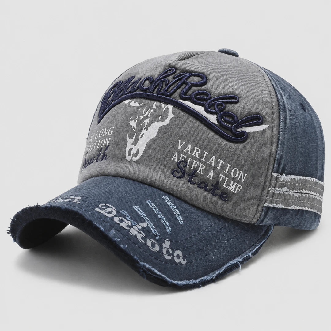 Josh & Tate - BlackRebel™ Distressed Cap - A distressed baseball cap with a gray front panel and a dark blue denim brim and side panels. The front features embroidered text reading 'Black Rebel' in dark blue, a white cowboy silhouette, and white text reading 'Dakota' and 'VARIATION AFTER A TLFMP State'.